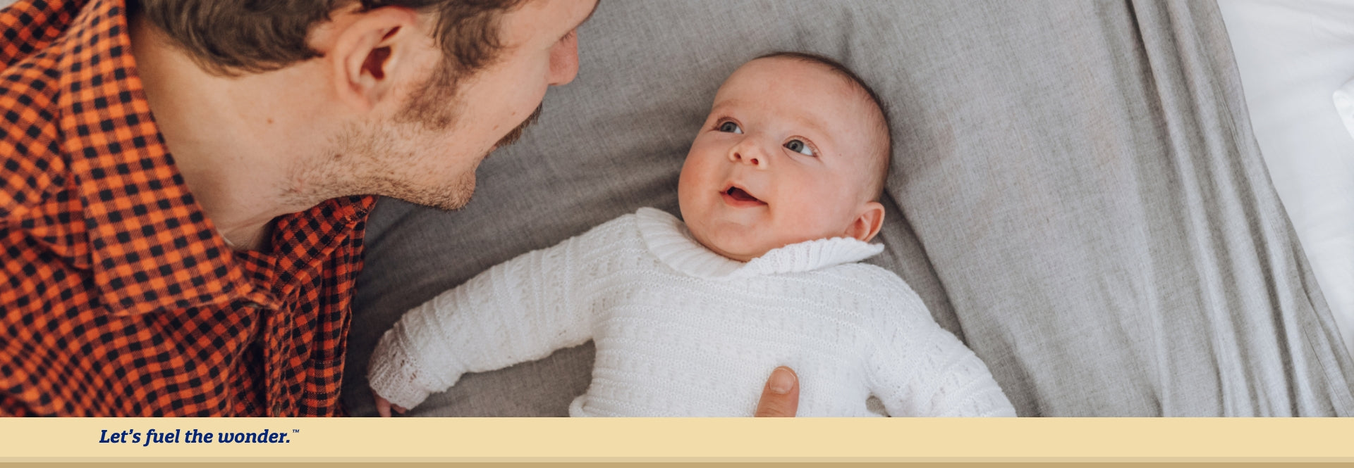 Baby laying on their back on a large bed with arms stretched out, staring up at baby's father as they look down towards baby and support baby's side with their right hand