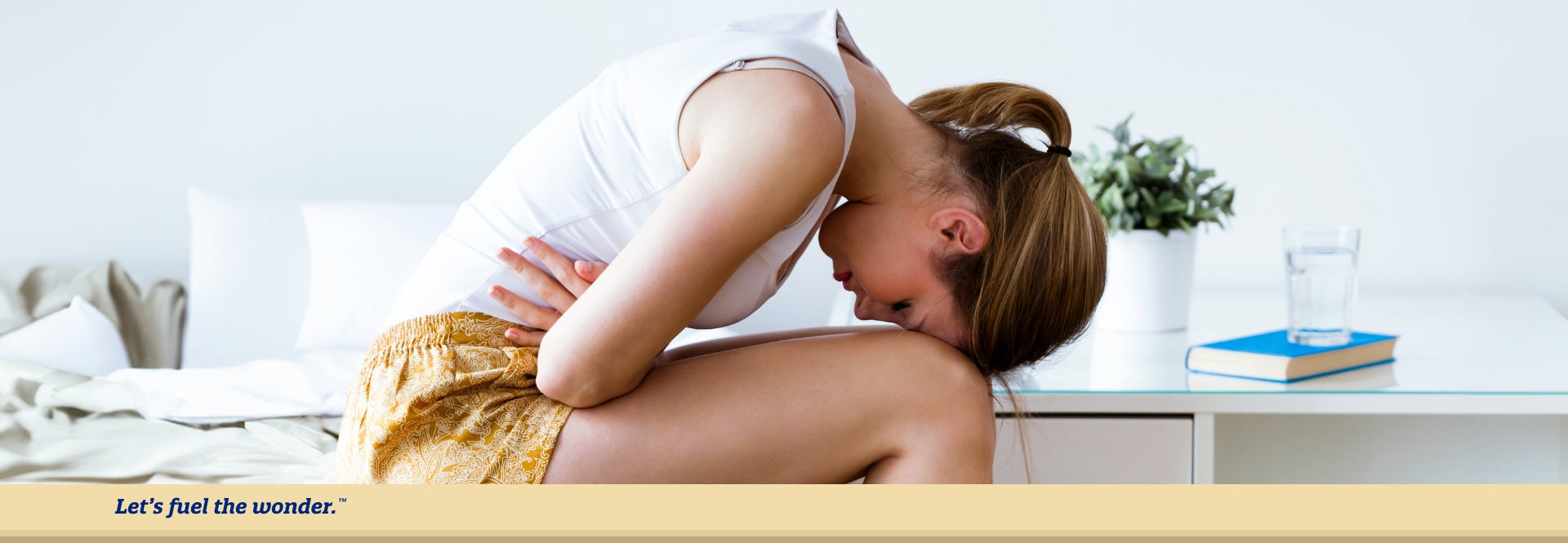 Woman with eyes closed sitting down on bed, leaned over with head touching their knees while clenching their stomach in mild pain