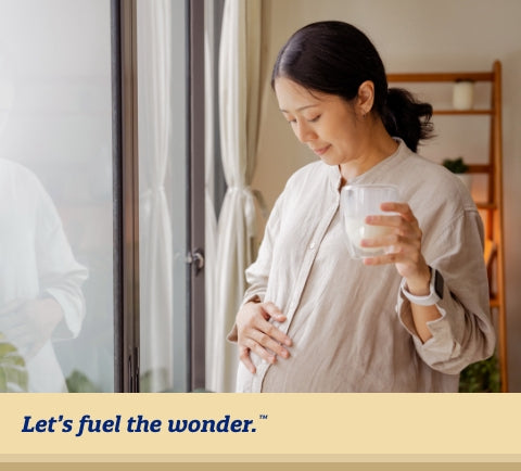 Pregnant woman standing by window looking down at bellow while holding a glass of milk in left hand and rubbing pregnant belly with right hand